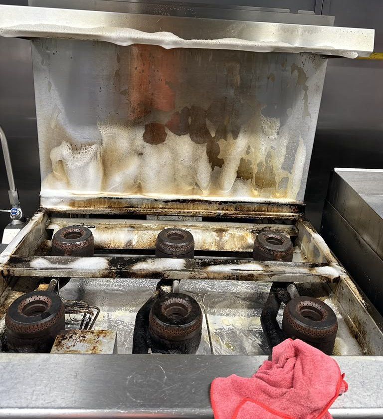 Commercial kitchen range being degreased, foam lifting built-up grease from the backsplash and burners