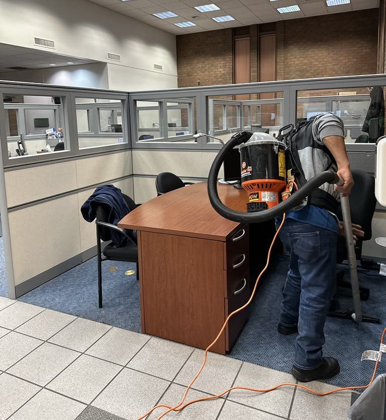 Janitor using a backpack vacuum to clean carpet in an office cubicle